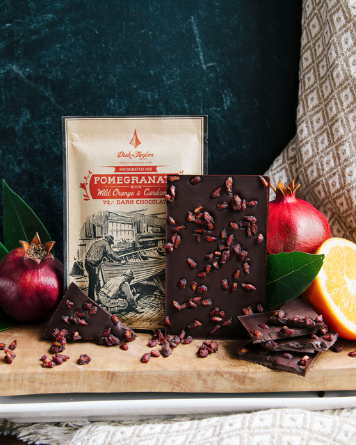 Pomegranate with Wild Orange & Cardamom package dark chocolate with full bar, stack of chocolate and flanked with ripe pomegranate/orange fruit
