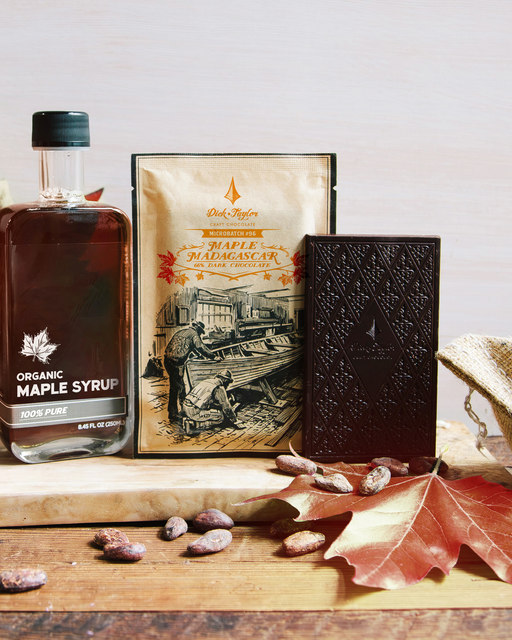 Maple Madagascar bar in package, dark chocolate bar, bottle of maple syrup on a cutting board. Scattered cocoa beans and a red maple leaf.